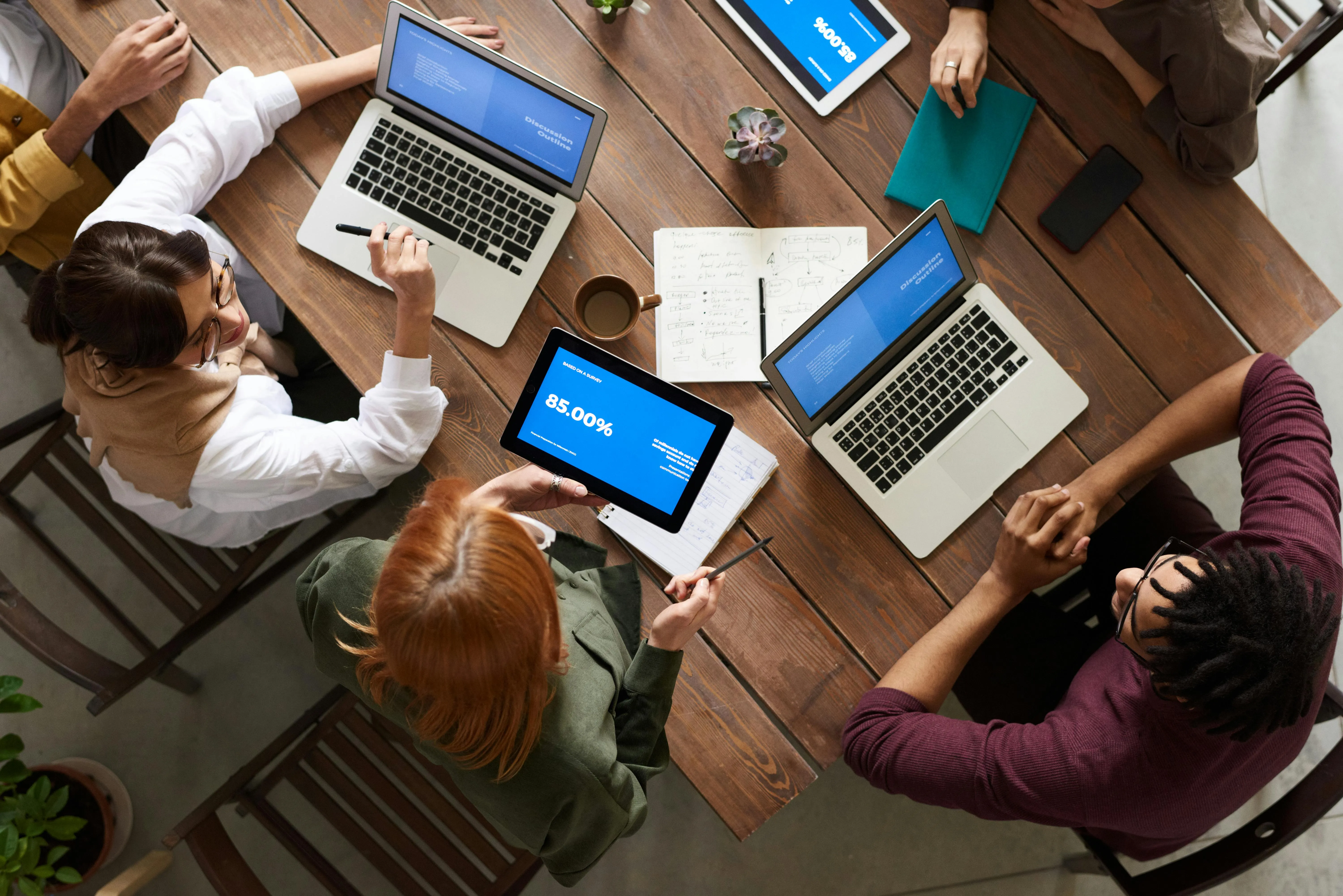 #FotoDescrição: Quatro pessoas em volta de uma mesa de madeira em uma reunião. Todos têm notebooks ou tablets abertos com a tela mostrando a palavra "BALANCE". Há cadernos, anotações, café e canetas sobre a mesa, sugerindo uma sessão de planejamento ou trabalho colaborativo.