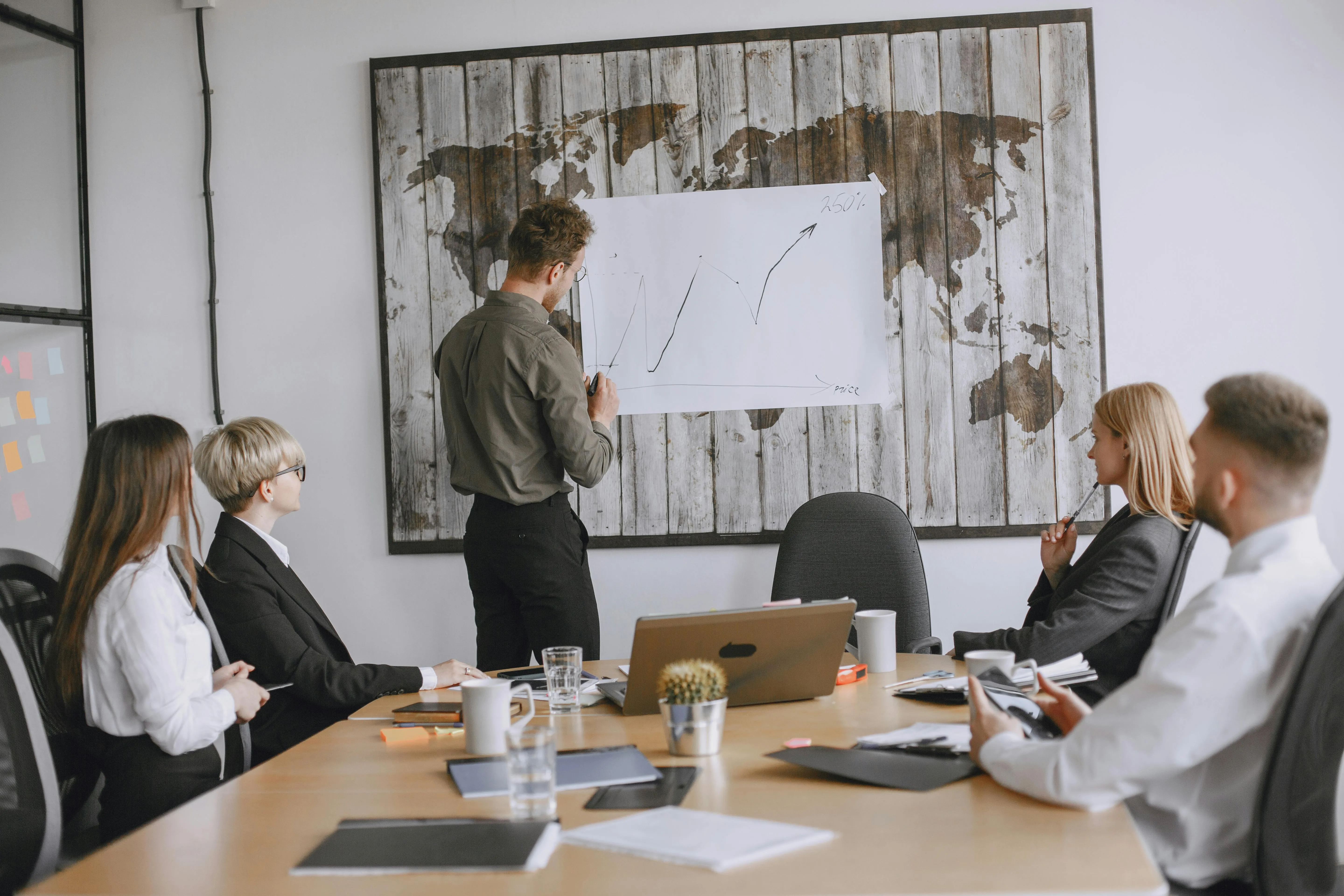 #FotoDescrição: Homem de camisa preta apresentando um gráfico de crescimento em um painel para uma equipe em sala de reunião com decoração de mapa-múndi.