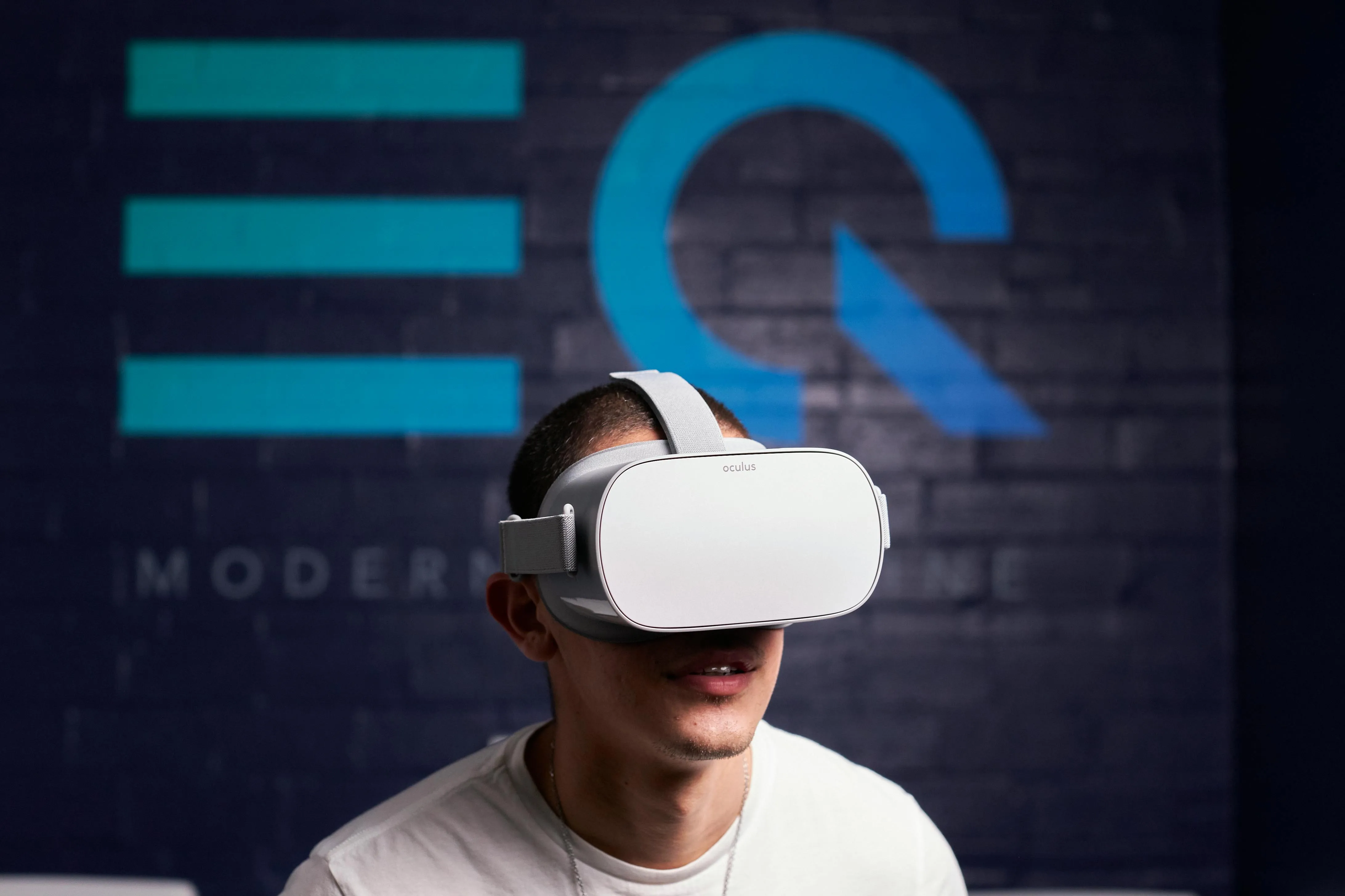 #FotoDescrição Homem utilizando um óculos de realidade virtual com expressão de surpresa ou curiosidade, em frente a uma parede com logotipo moderno, representando experiências imersivas e tecnológicas.