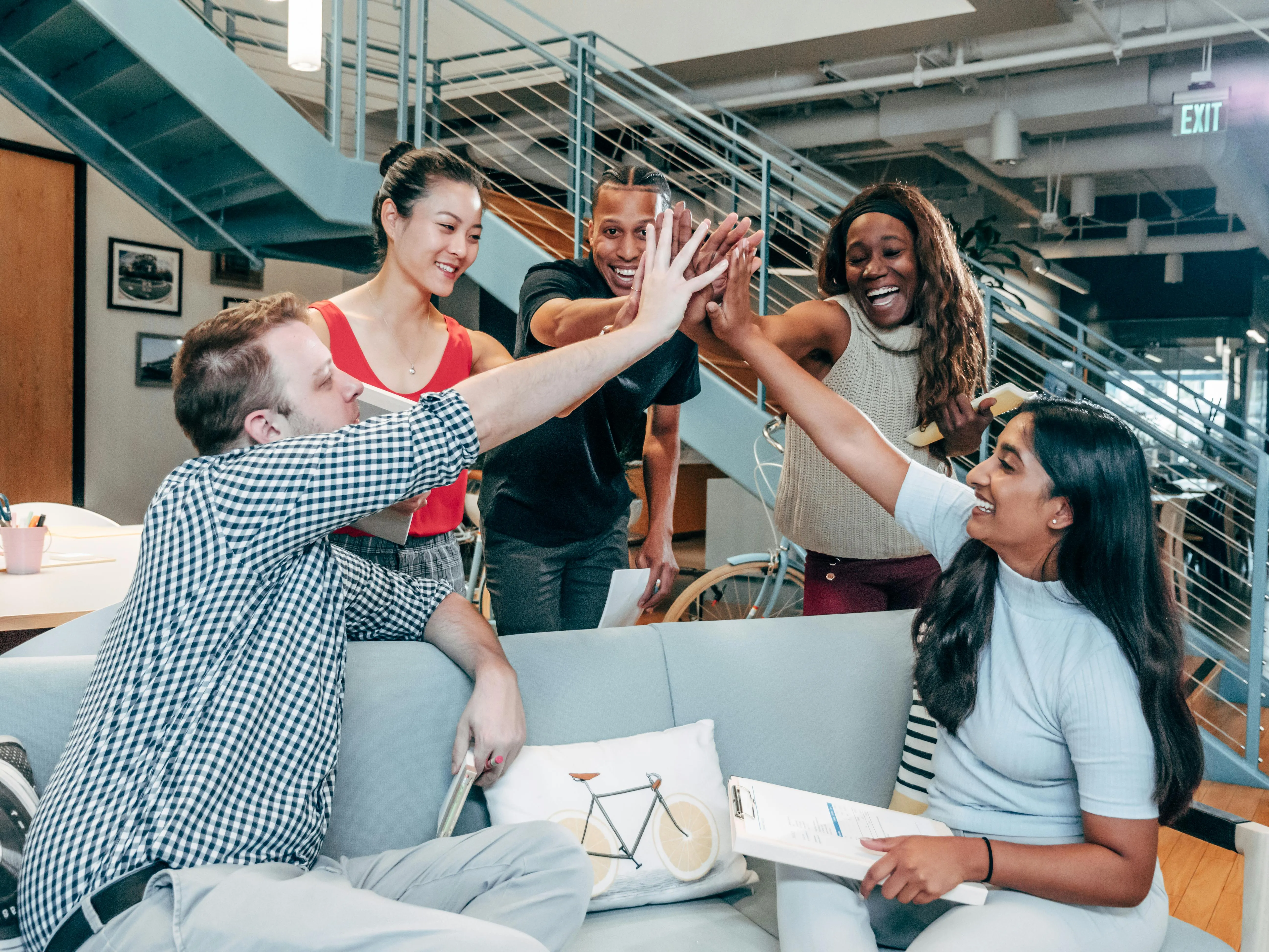 #FotoDescrição: Grupo de cinco colegas de trabalho sorrindo e celebrando juntos com um high-five em um ambiente de escritório moderno.