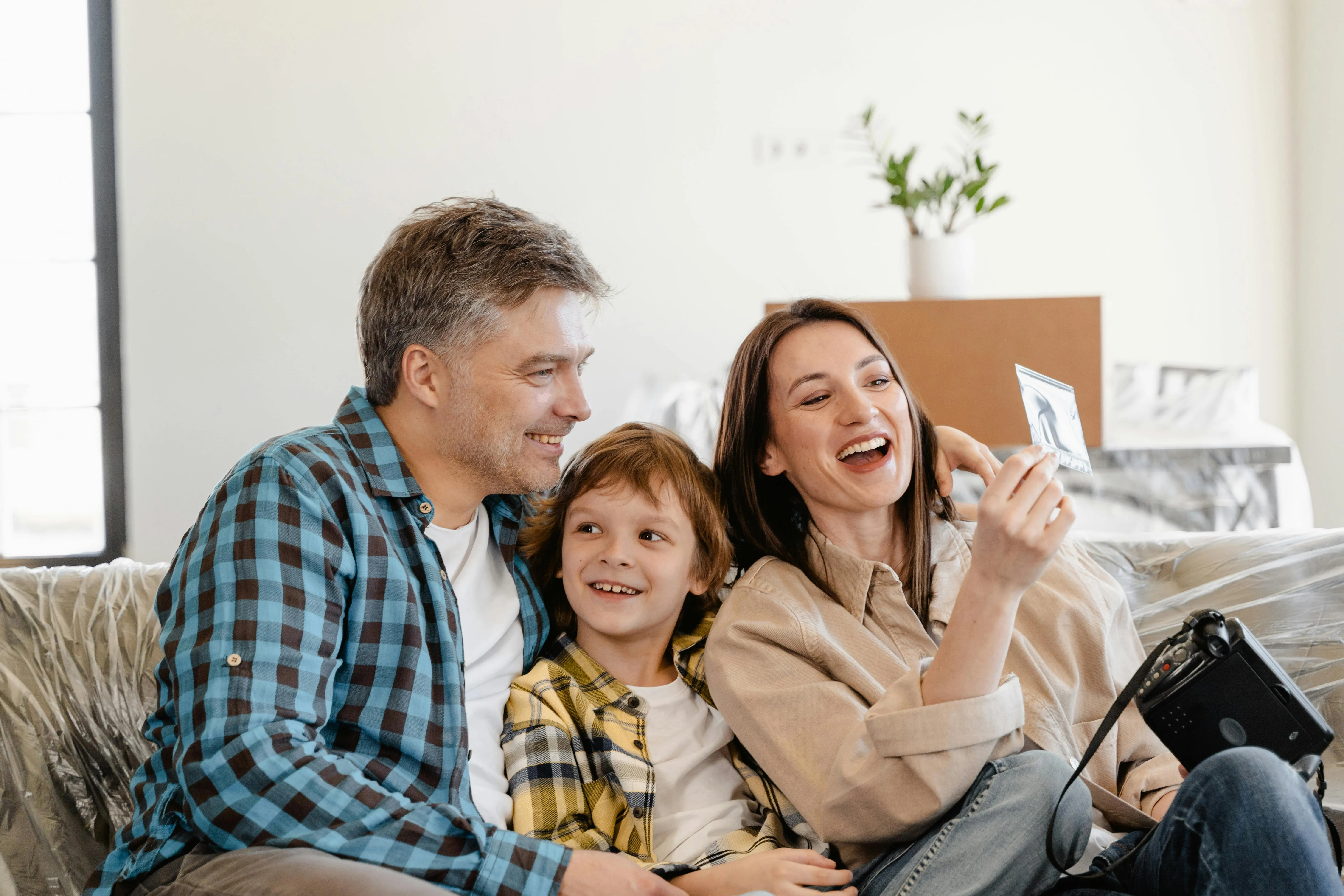 #FotoDescrição Pai, mãe e filho sorrindo sentados em um sofá, segurando uma fotografia impressa e uma câmera. Ao fundo, caixas e objetos sugerem um momento de mudança ou celebração familiar.