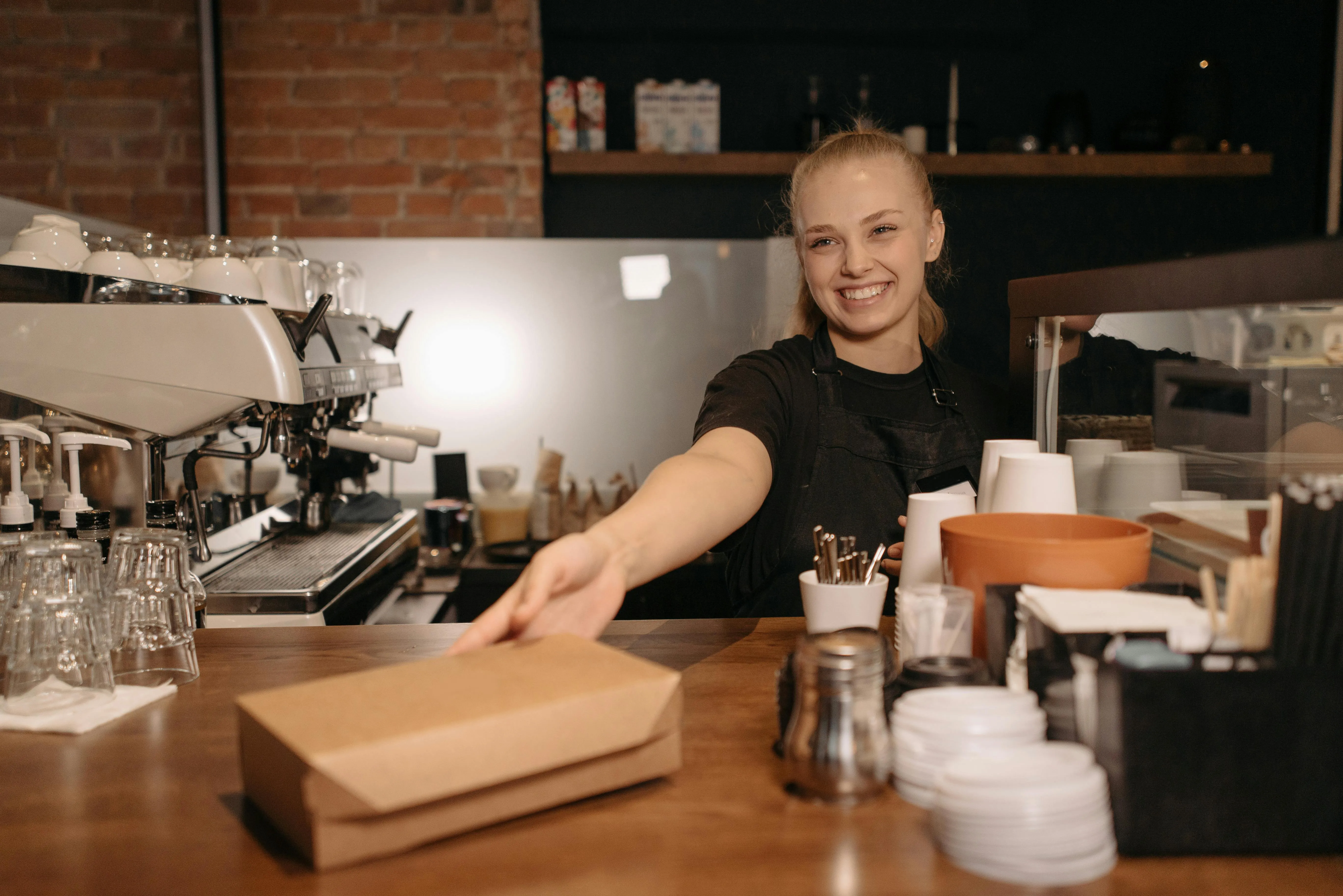 #FotoDescrição Jovem atendente loira sorrindo atrás do balcão de uma cafeteria, estendendo uma embalagem de papel kraft para o cliente.