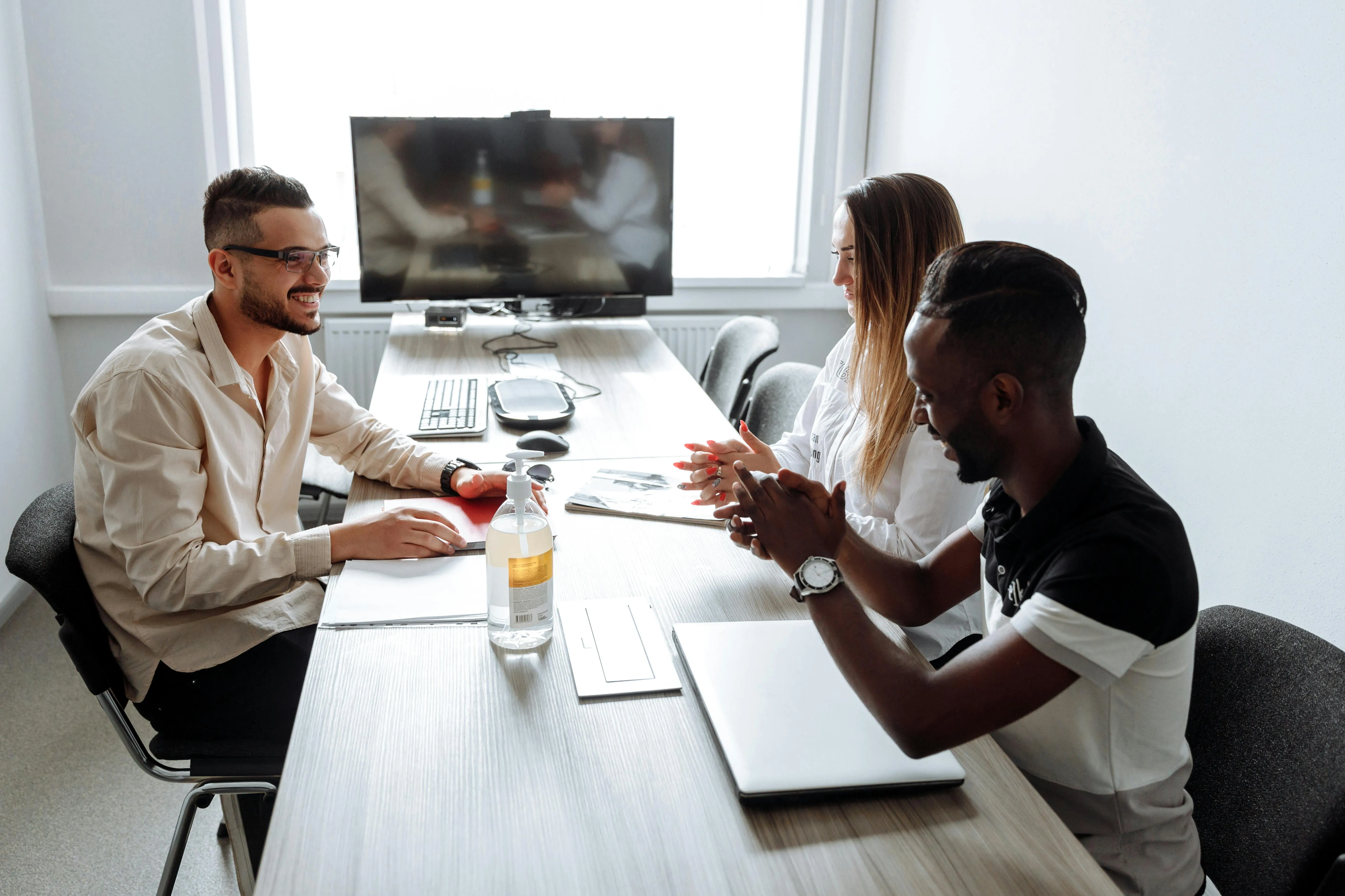 #FotoDescrição Três jovens adultos reunidos em uma sala de reunião moderna, conversando e sorrindo em torno de uma mesa com notebooks e garrafa de água, simbolizando colaboração e engajamento em projetos educacionais ou corporativos.