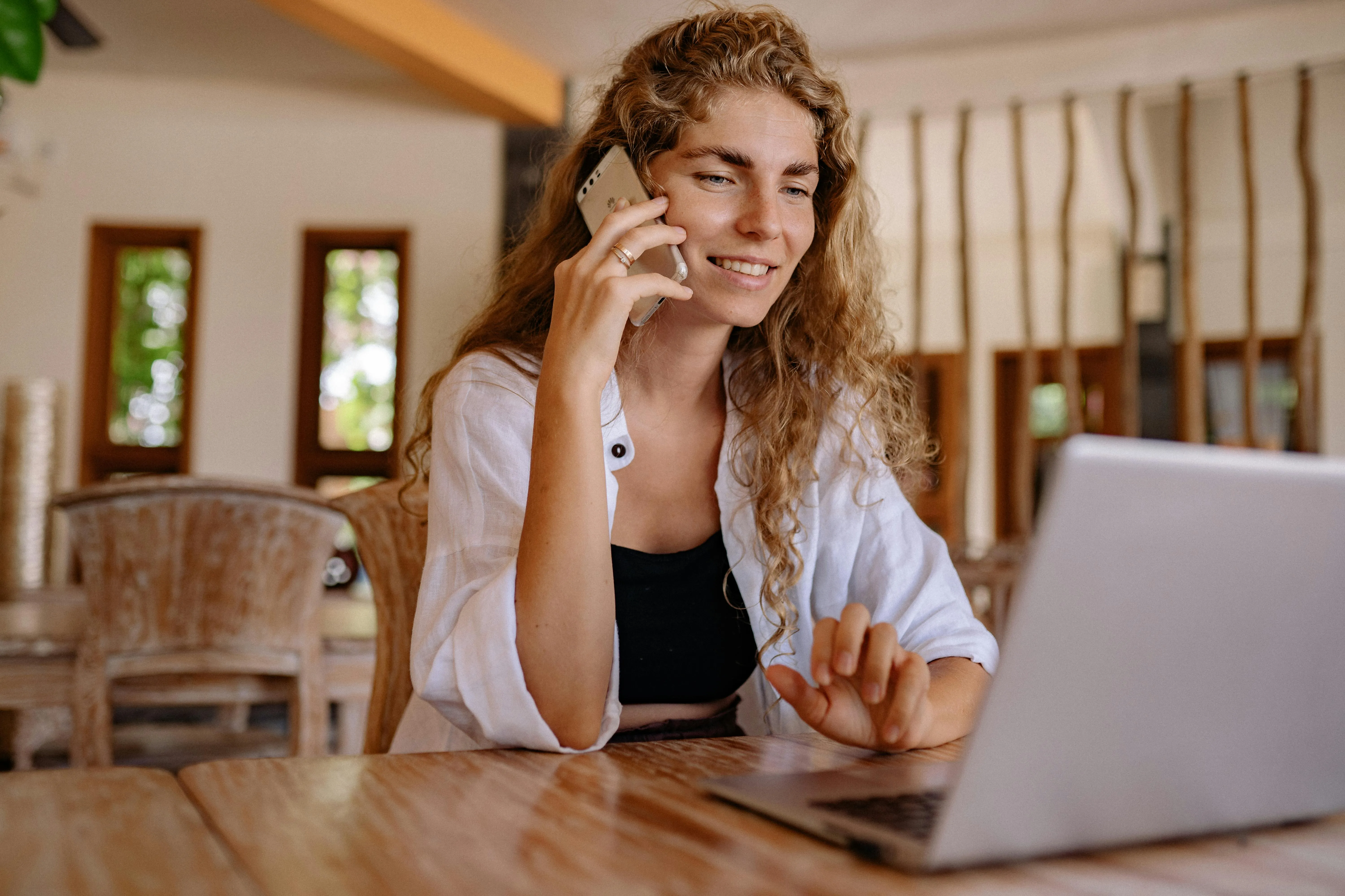 Mulher sorrindo ao falar ao telefone e usar laptop em mesa de madeira.