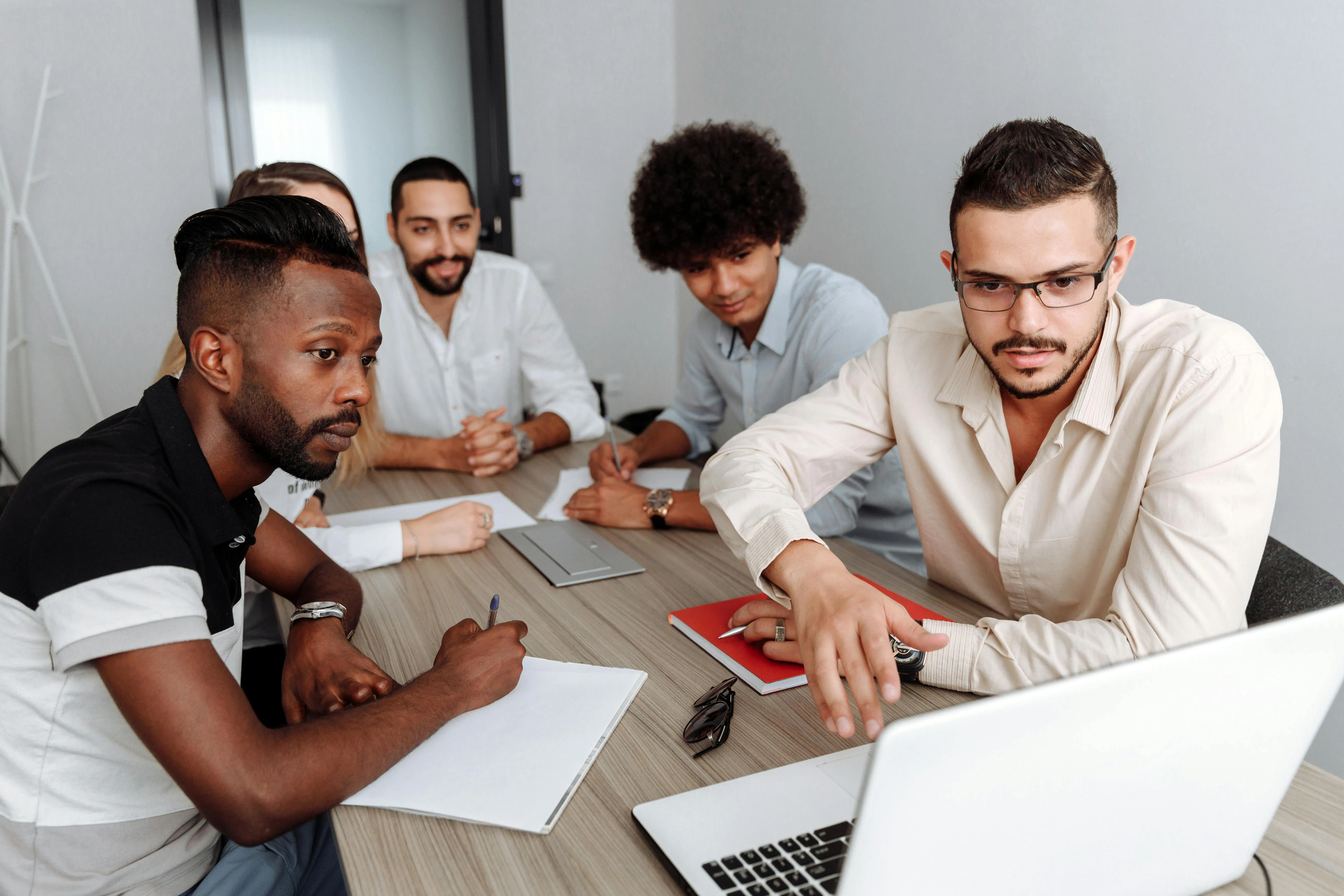 #FotoDescrição Grupo de cinco pessoas reunidas em torno de uma mesa, discutindo estratégias com foco em um notebook, representando o planejamento de produtos digitais em equipe.