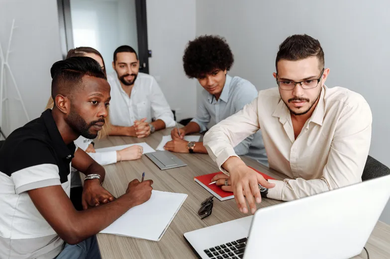 #FotoDescrição Grupo de cinco pessoas reunidas em torno de uma mesa, discutindo estratégias com foco em um notebook, representando o planejamento de produtos digitais em equipe.