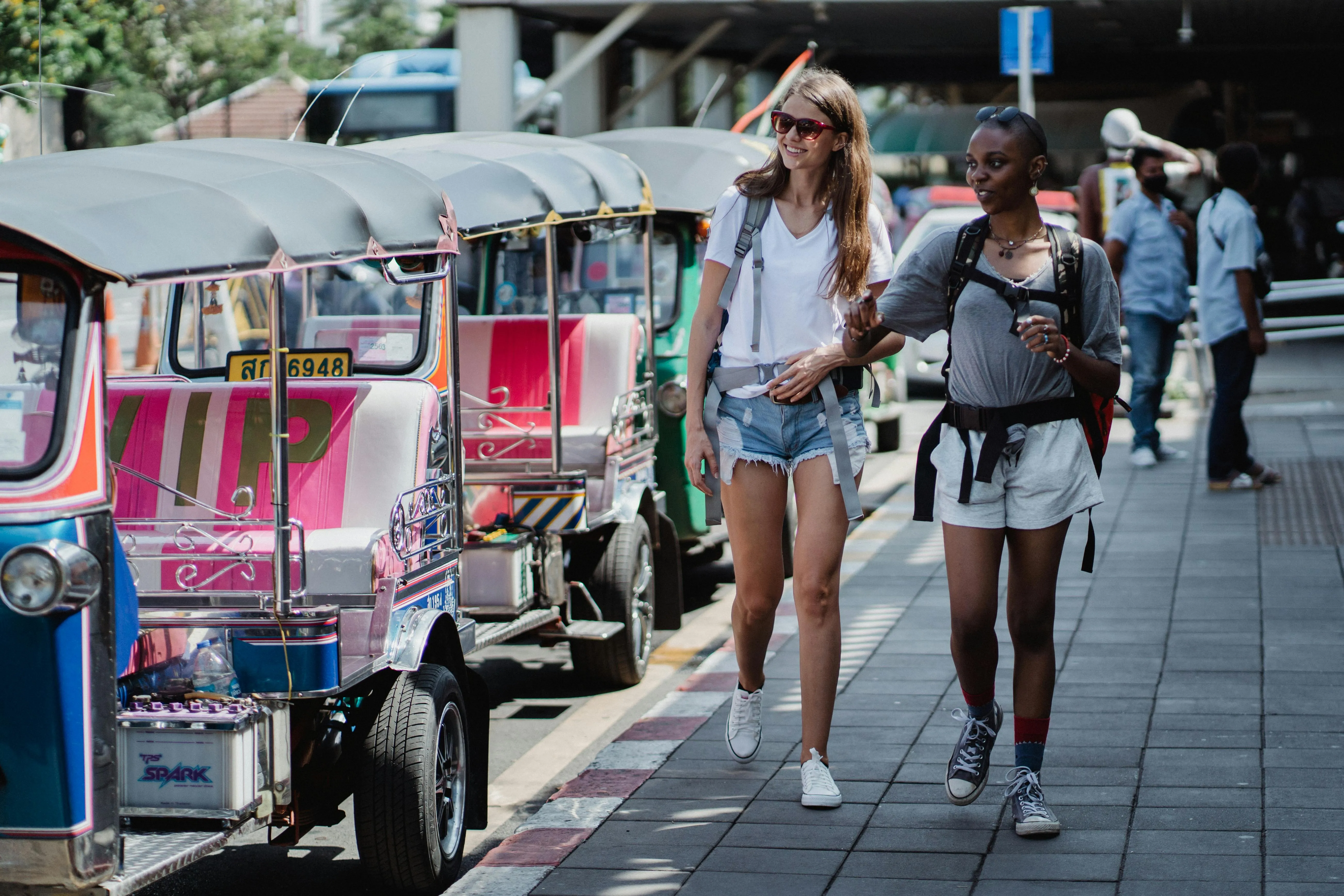 Duas viajantes caminham por rua urbana ao lado de tuk-tuks coloridos