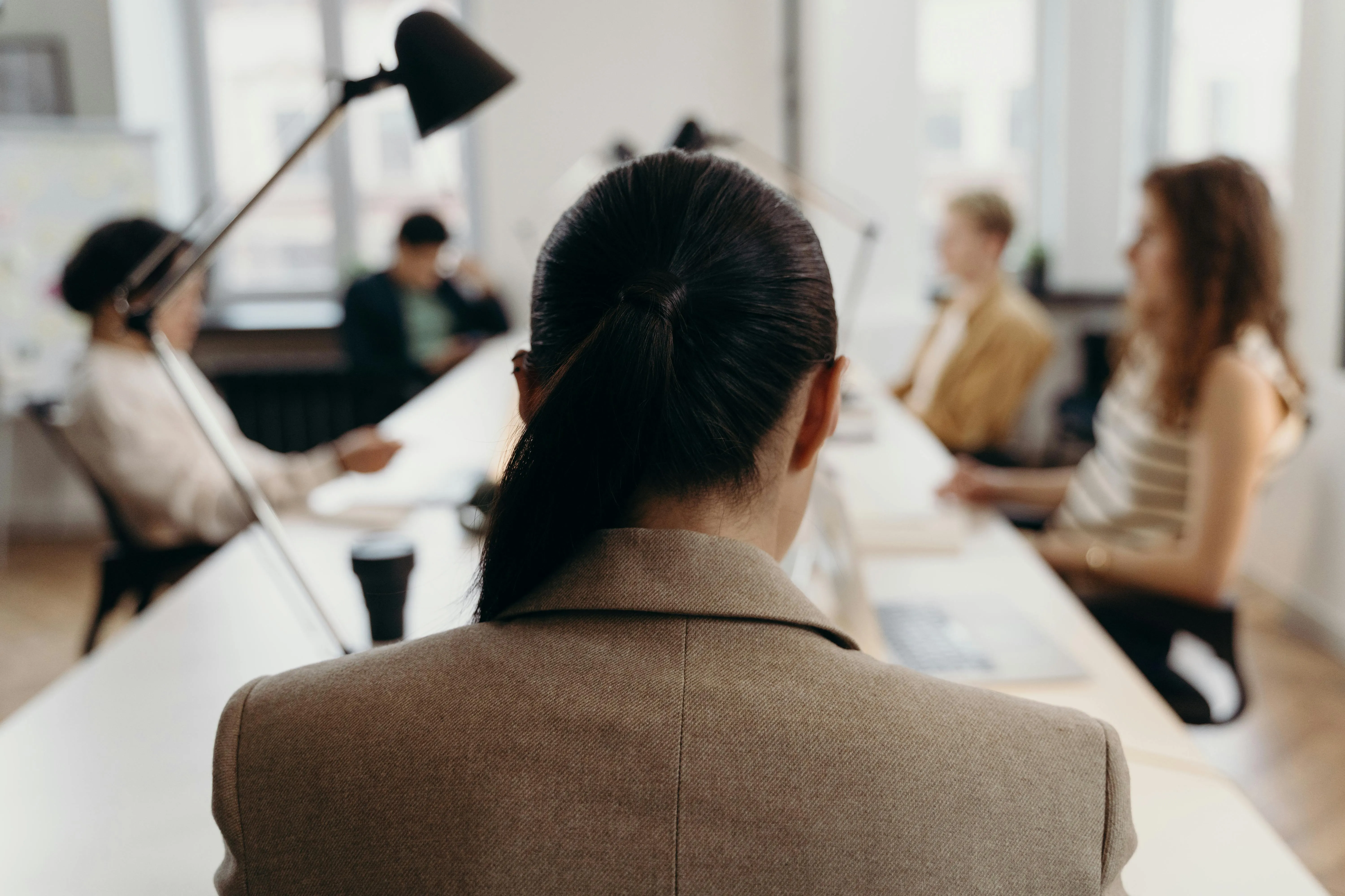 #FotoDescrição Vista traseira de uma mulher de cabelo preso, sentada à mesa em uma sala de reunião moderna, com colegas ao fundo, simbolizando foco, liderança e tomada de decisão em equipe.