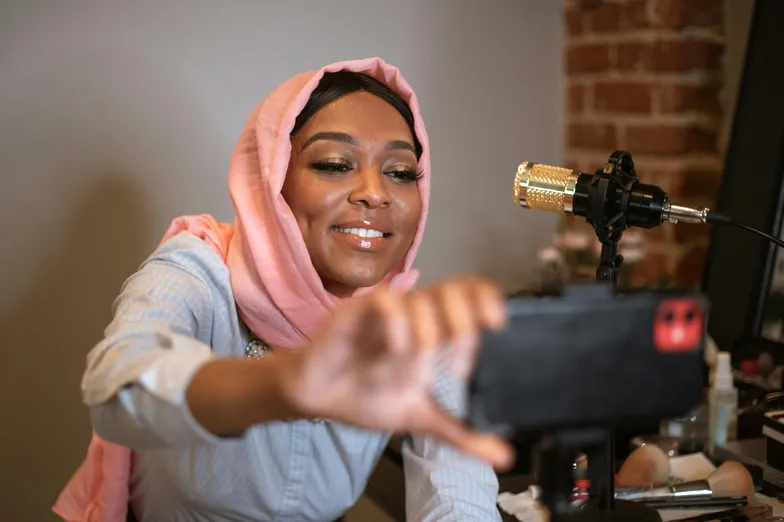 Mulher sorridente com hijab rosa, tirando selfie em um estúdio de gravação com microfone dourado ao fundo.