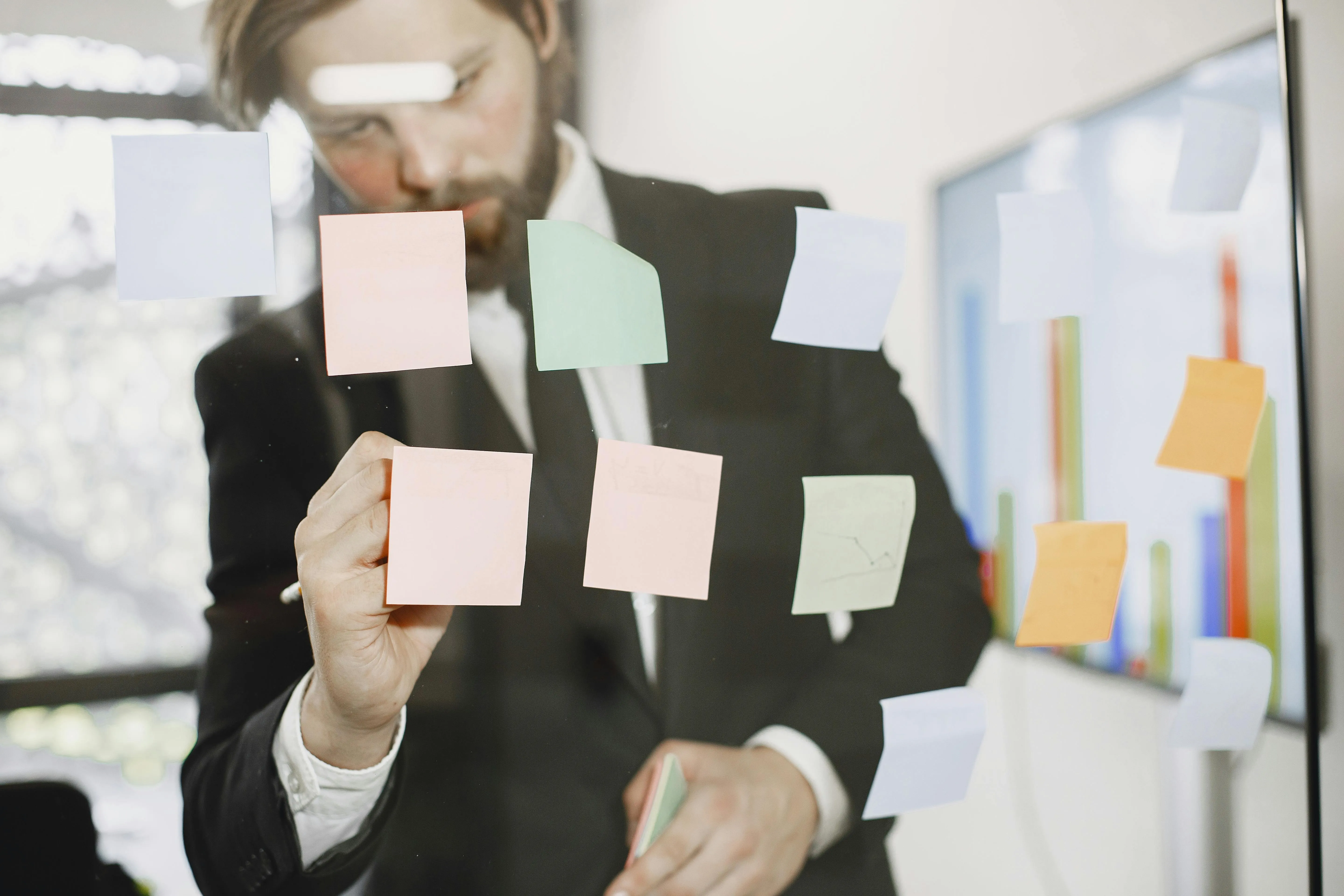 #FotoDescrição Homem de terno colando post-its coloridos em uma parede de vidro transparente em ambiente corporativo, representando planejamento estratégico, organização e estruturação de ideias.