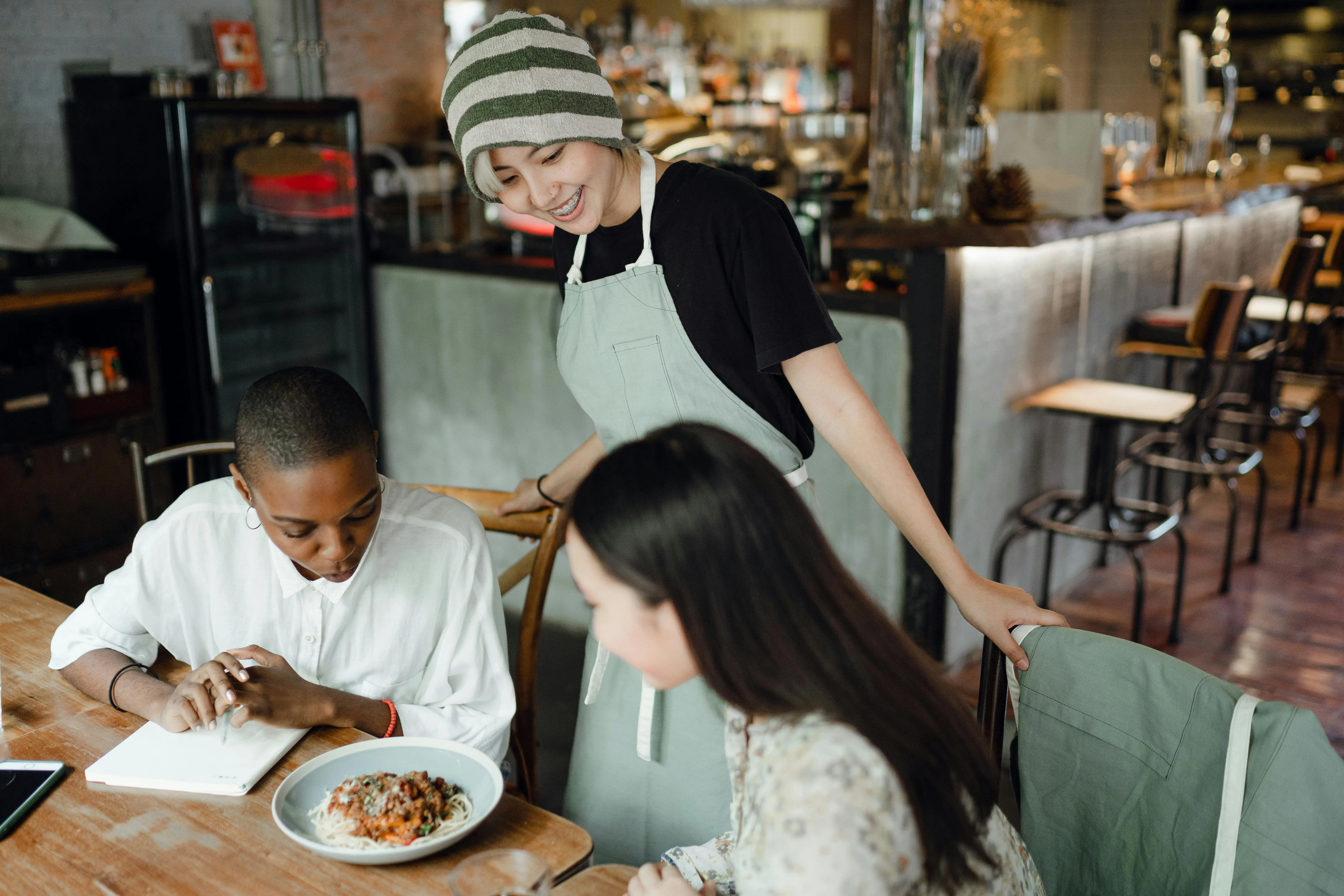 #FotoDescrição: Garçonete sorrindo enquanto atende dois clientes sentados à mesa em um restaurante moderno, com um prato servido à frente.
