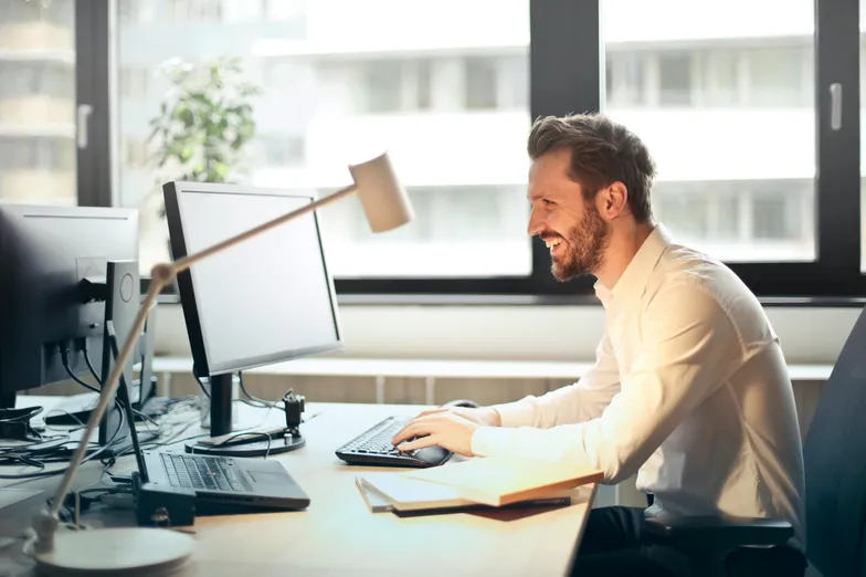 #FotoDescrição Homem sorridente trabalhando em frente a dois monitores em um escritório moderno com luz natural, aparentando estar satisfeito ou engajado com o que vê na tela.